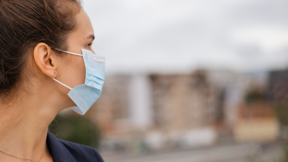 Side profile of a woman wearing a blue surgical mask looking into the distance with blurred city buildings in the background.