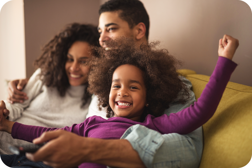 Smiling young girl in purple shirt sitting on a couch with her parents, the father holding a remote control.